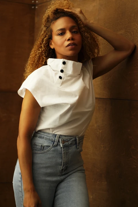 Daisi Jo — in a white button-neck top and jeans with one hand in her hair