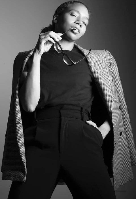 Daisi Jo in a checked blazer and black trousers, holding glasses near her lips